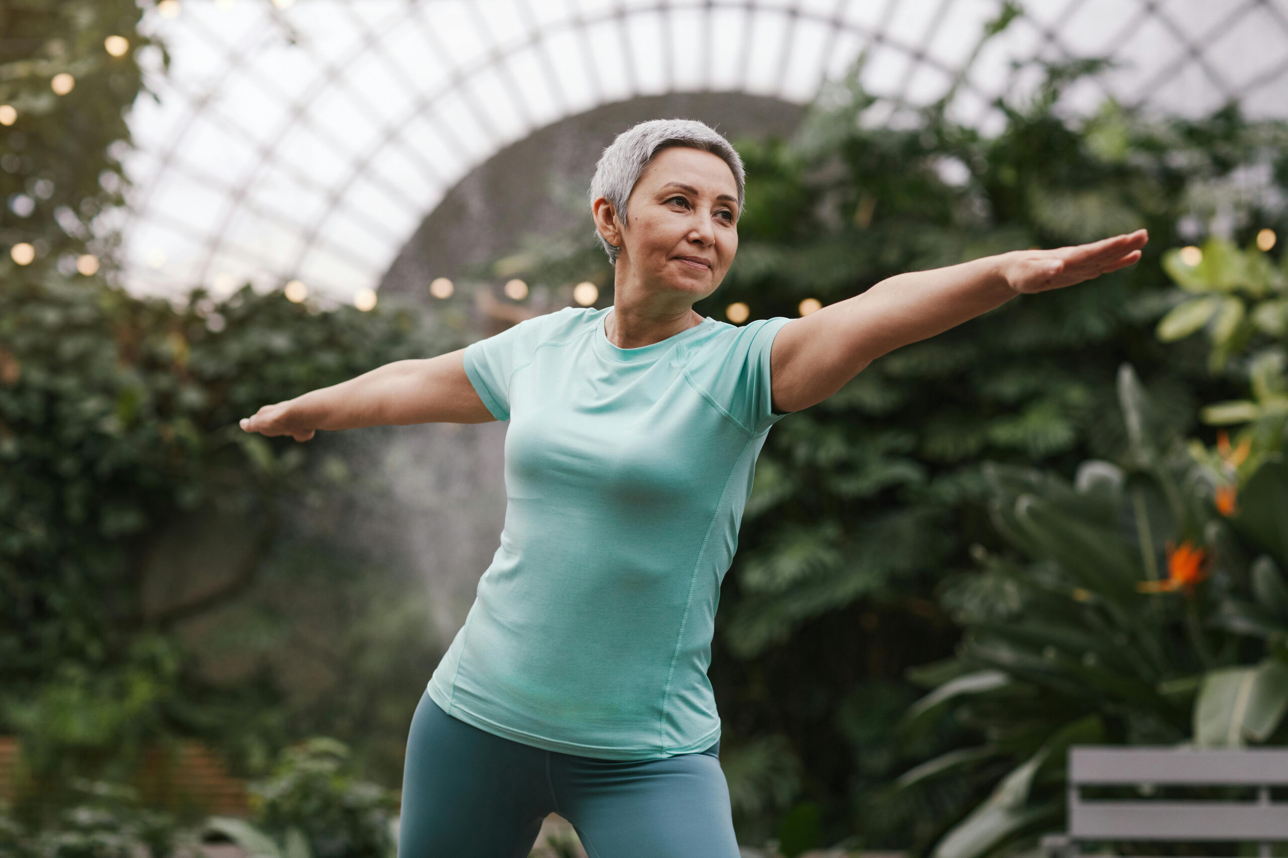 Active senior doing yoga stands in warrior 2 pose, amid a room full of plants